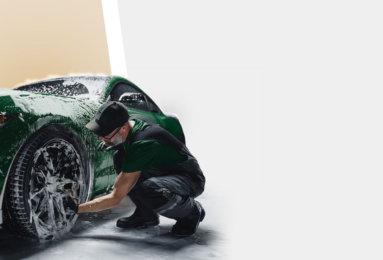 Mann in schwarzer Kappe und grüner Arbeitskleidung wäscht grünes Auto und reinigt Rad mit Schwamm